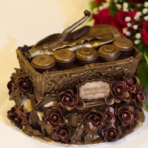 Coklat tepak sireh bersalut gangsa, dengan lima cembul bertutup di atasnya, bunga ros coklat gelap di sekeliling dindingnya dan plak Chocolate Chapter di hadapan, di sebelah jambangan ros merah